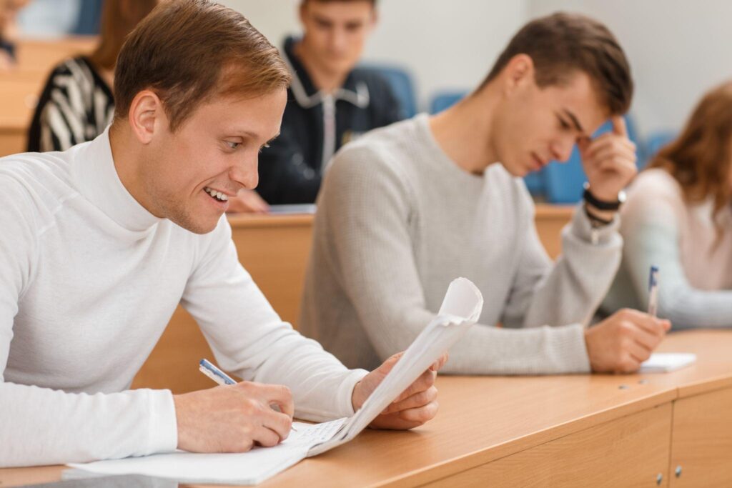 male student sitting at table, smiling and copying notes at university