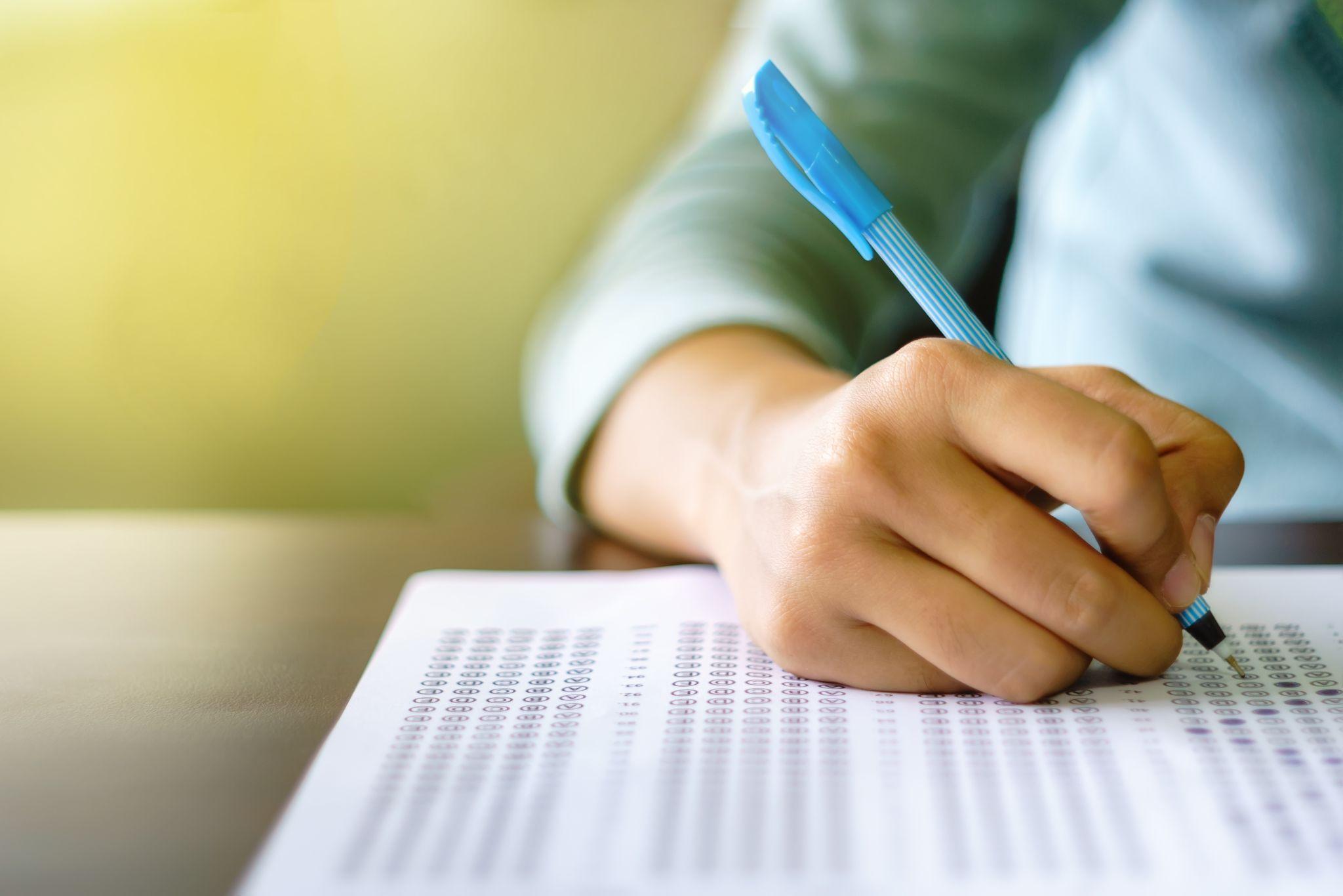 Close up of high school or university student holding a pen writing on answer sheet paper in examination room.